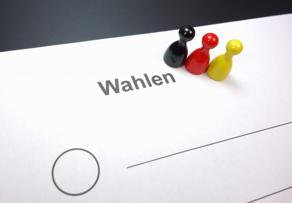 Drei Spielfiguren in Deutschlandfarben stehen auf einem Wahlzettel